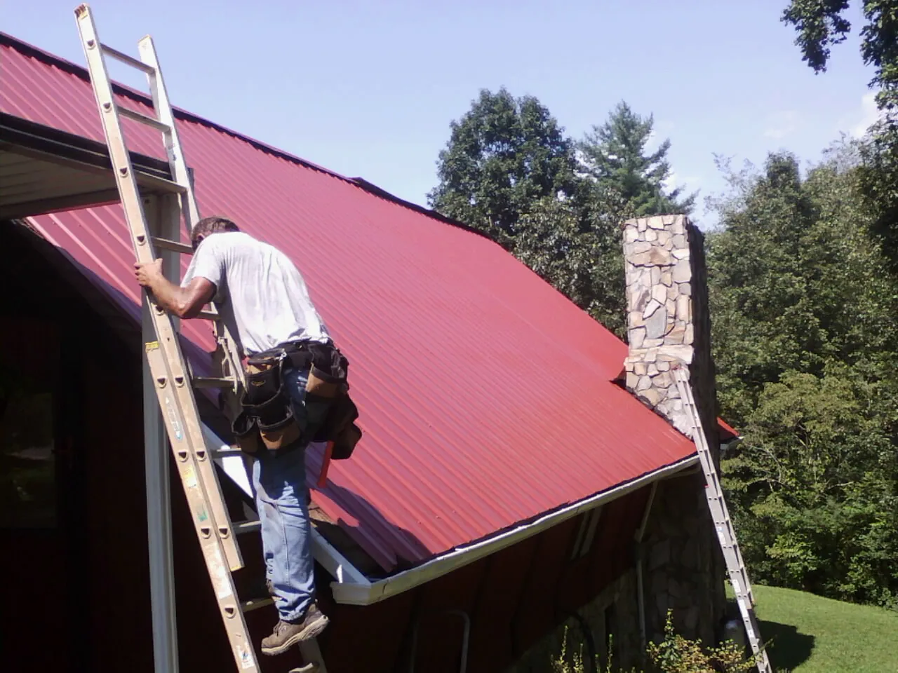 Roofer on a ladder beside a red metal roof and stone chimney — Soffit & Fascia Repair by Roofing Brothers in Platina